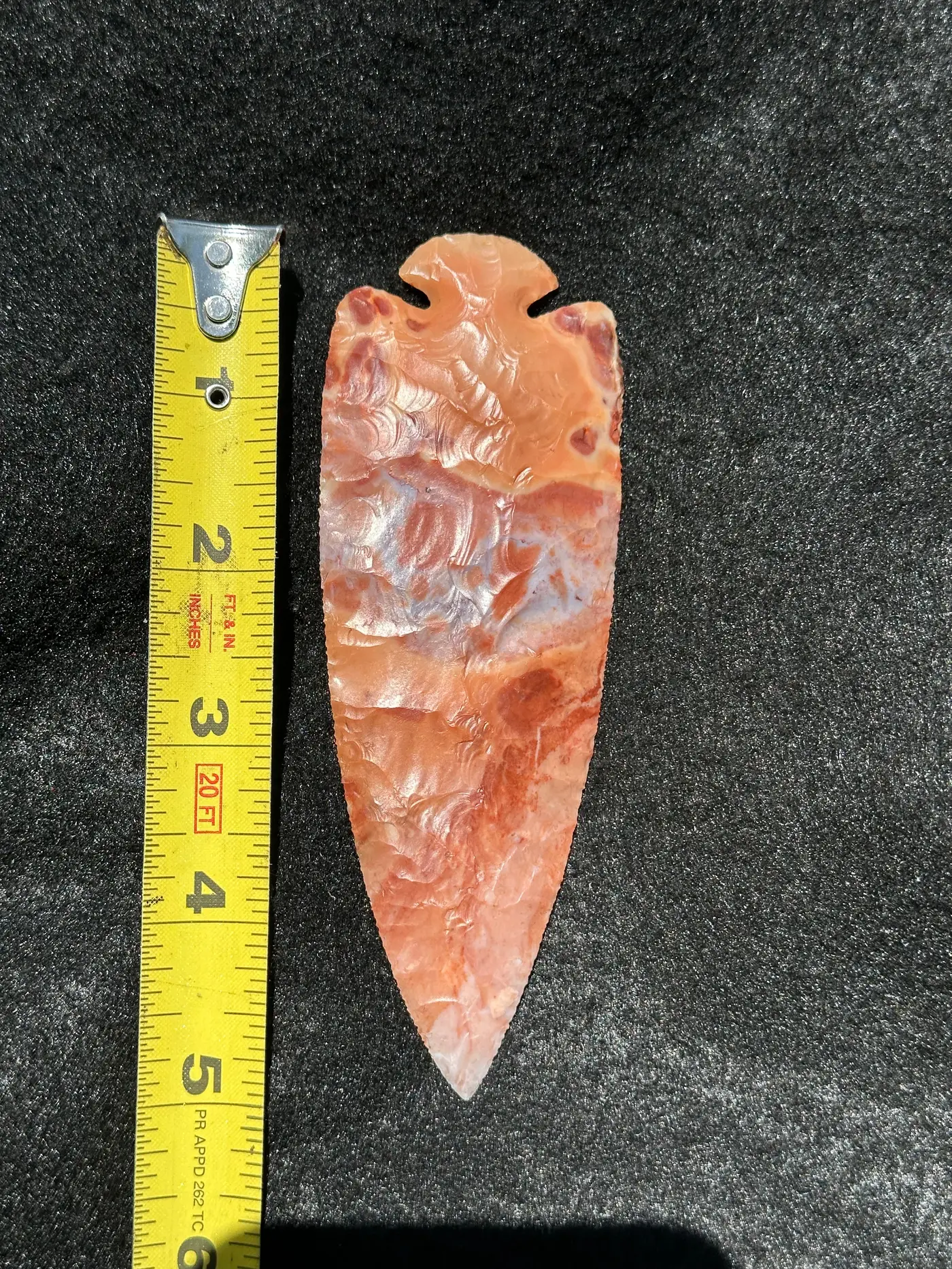 Front of a long red-orange dovetail flint point with marbled cream and gray areas beside a tape measure.