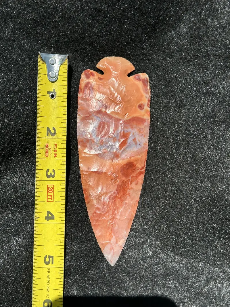 Front of a long red-orange dovetail flint point with marbled cream and gray areas beside a tape measure.