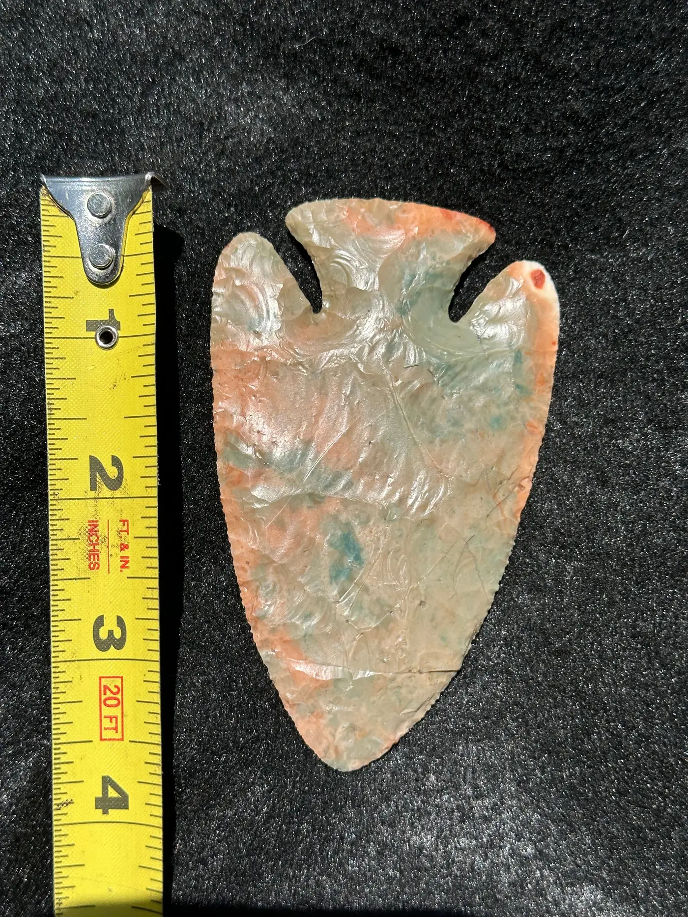 Pale cream and gray deep-notched flint point with peach and blue-green markings beside a tape measure.